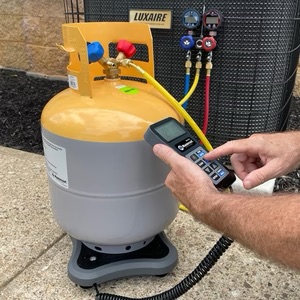 Technician holding a charging-scale remote next to a refrigerant cylinder standing on a scale in front of the outdoor unit, with gauges and hoses behind